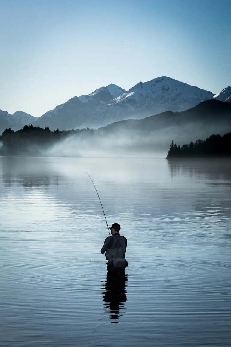 Pescador con mosca de pie en un lago rodeado de montañas nevadas